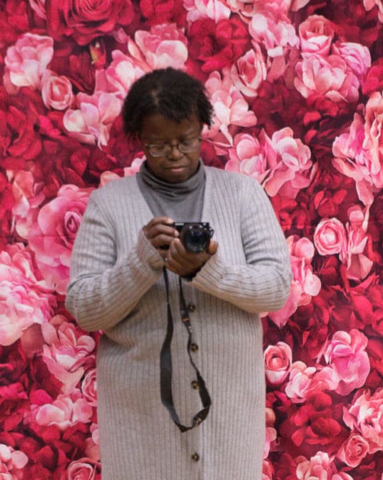 A woman holding a camera in front of a floral background.